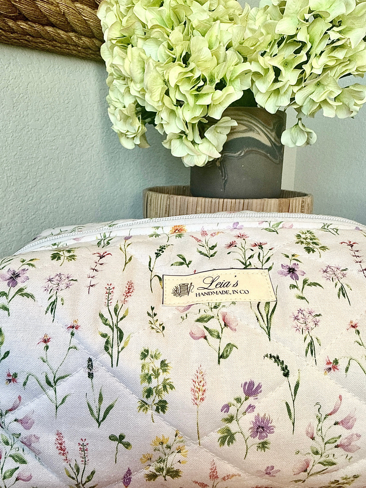 A quilted fabric zippered pouch with a delicate multicolor wildflower print on a cream background, featuring a visible 'Lena's Handmade Co.' label.