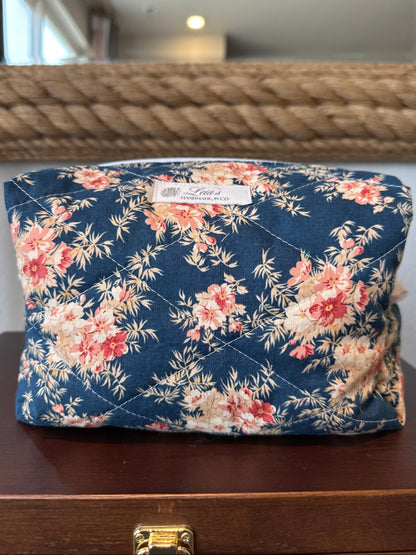 A quilted cosmetic pouch with a navy blue floral print featuring pink and red flowers, resting on a dark wooden surface in front of a mirror.