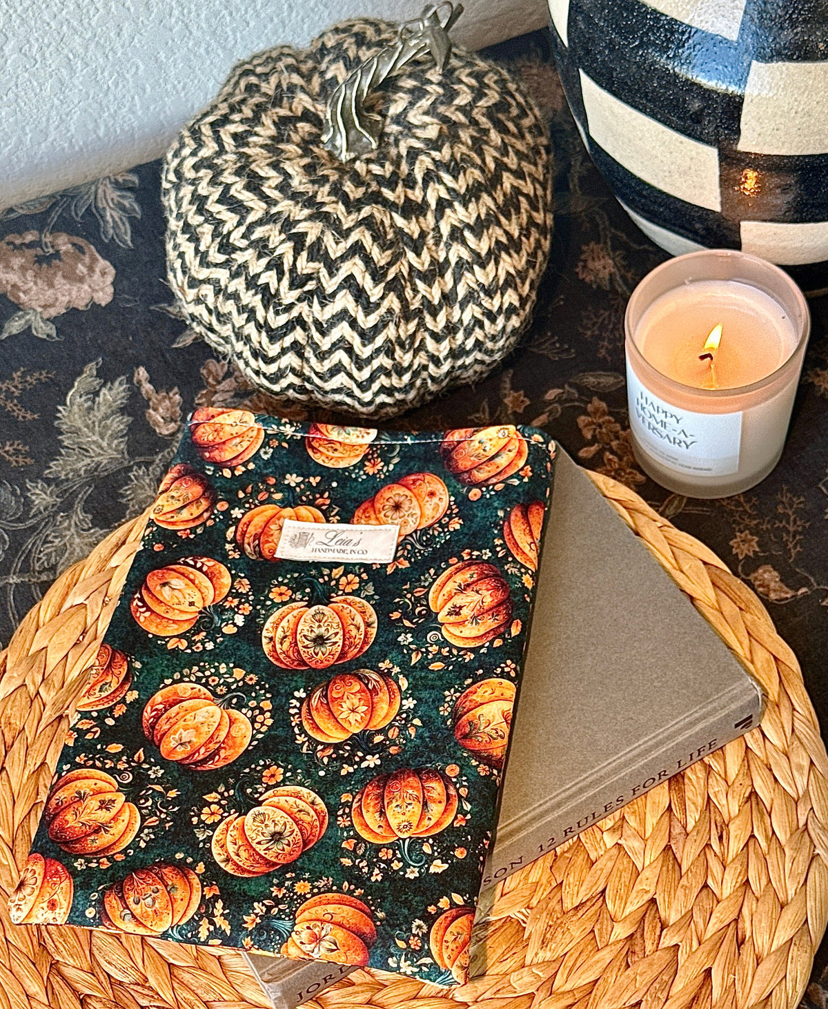 (angled flat lay, book partially visible beneath sleeve)
Handmade quilted book sleeve in Gilded Gourd pumpkin print resting on a wicker charger next to a candle and decorative pumpkin