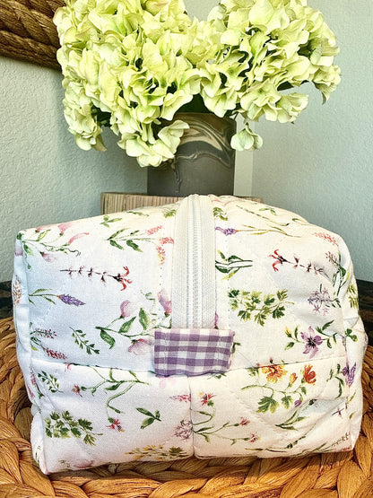 A quilted fabric cosmetic bag with a floral print and a purple gingham accent tab on the zipper, resting on a woven wicker tray with a vase of pale green hydrangeas in the background.
