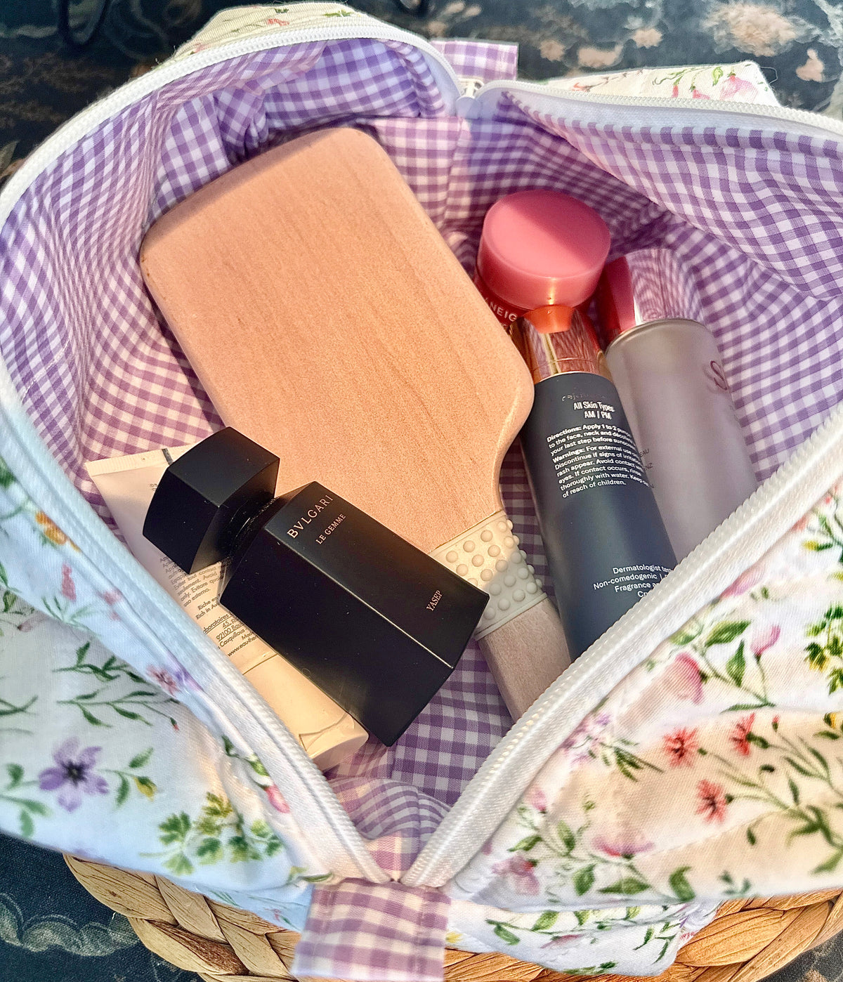A floral and gingham cosmetic bag filled with skincare and beauty products, including a black bottle, a pink-capped brush, and a wooden paddle brush, photographed from above.