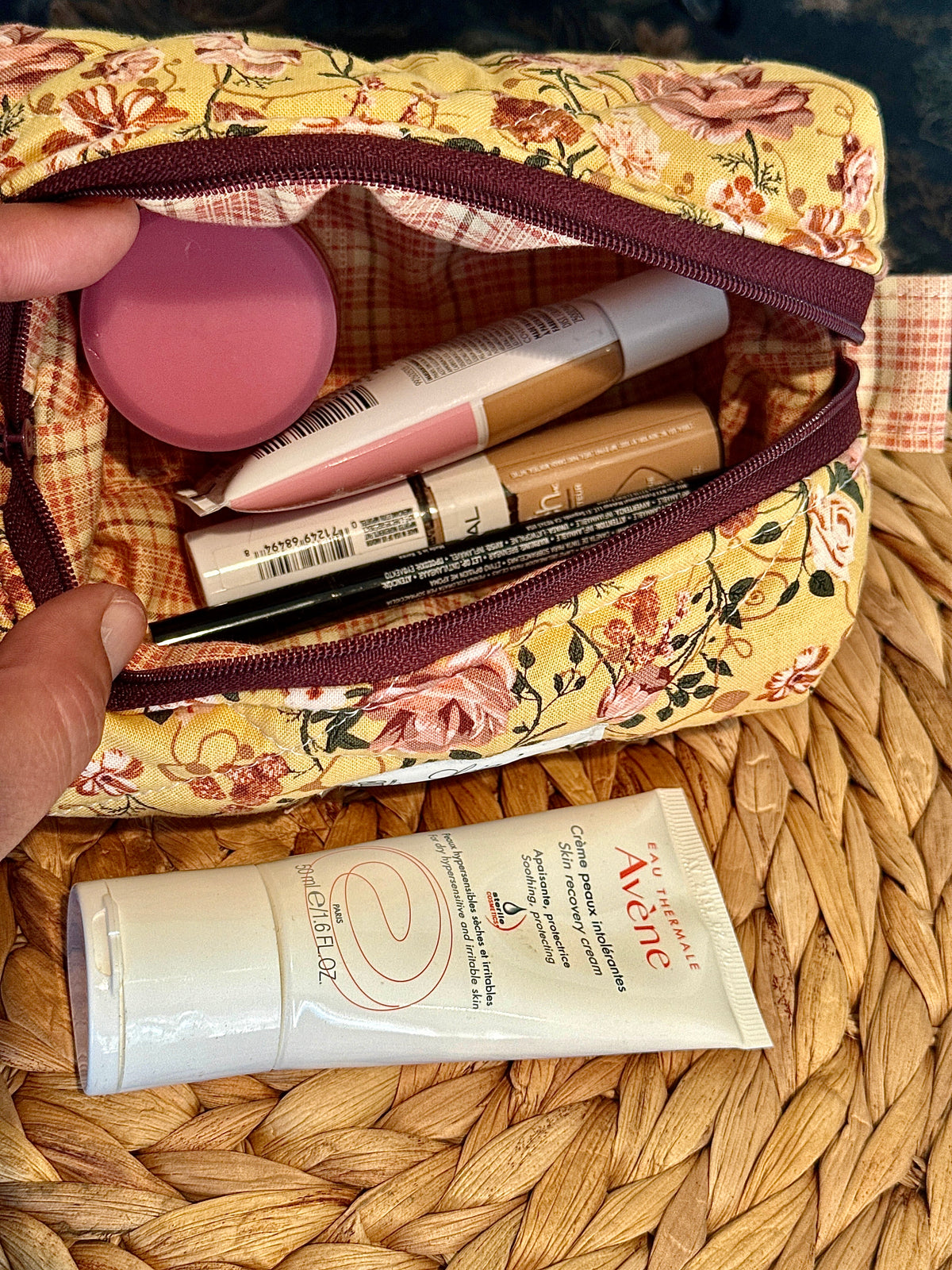 Open small floral boxy bag filled with makeup, showing its brown plaid lining and burgundy zipper