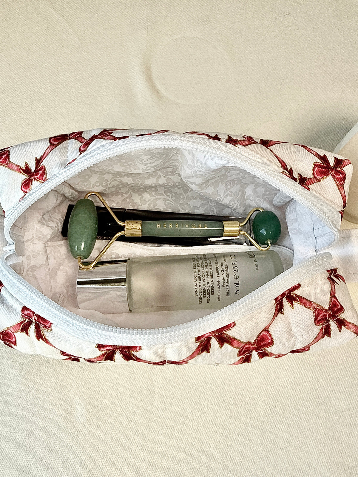 A white cosmetic pouch with a red bow pattern is shown open, revealing a jade roller and a skincare bottle inside.