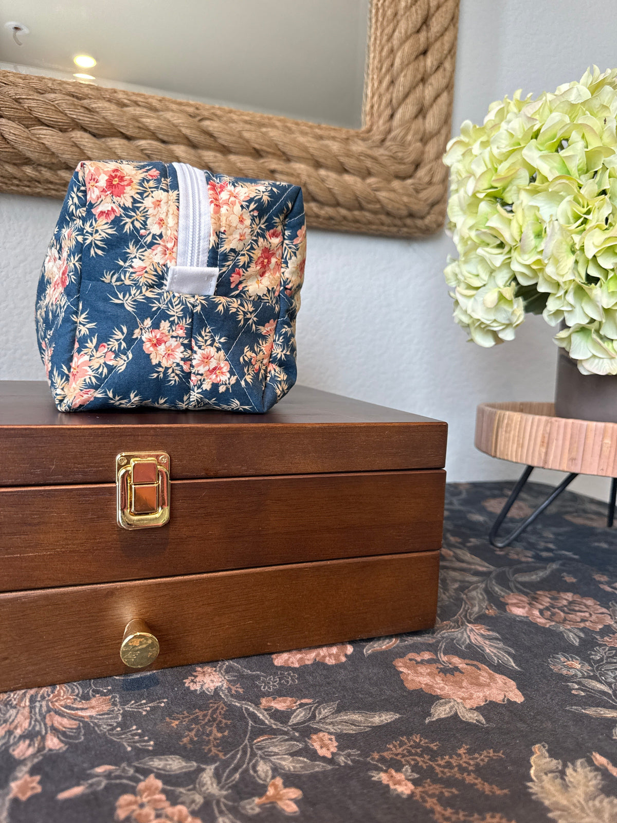 Side angle of the Ember & Indigo quilted boxy bag in navy floral fabric with a white zipper, showing its boxy shape and diamond quilting, on a wooden jewelry box.