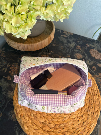 A fabric cosmetic pouch with a white floral print exterior and a purple gingham lining, shown open on a woven placemat with makeup items inside. A vase of pale green hydrangeas sits in the background on a patterned table surface.