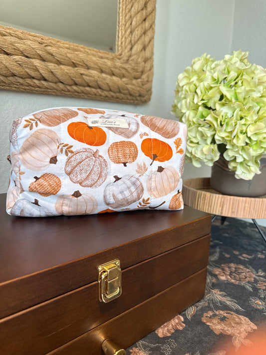 A fabric cosmetic pouch with an autumn pumpkin pattern sits on a dark wooden jewelry box in a styled home setting next to a potted green hydrangea.