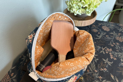 A fabric zippered makeup bag shown open, filled with a wooden paddle brush and cosmetics. The bag has a warm mustard-orange floral lining and a navy exterior with a white floral print.