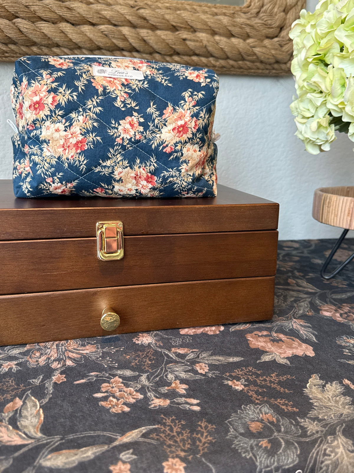 A navy blue floral makeup bag with red and pink flowers sits on top of a wooden jewelry box. The scene is styled with a matching floral rug, a rope-framed mirror, and a green hydrangea on a side table.
