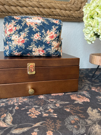 A navy blue floral makeup bag with red and pink flowers sits on top of a wooden jewelry box. The scene is styled with a matching floral rug, a rope-framed mirror, and a green hydrangea on a side table.