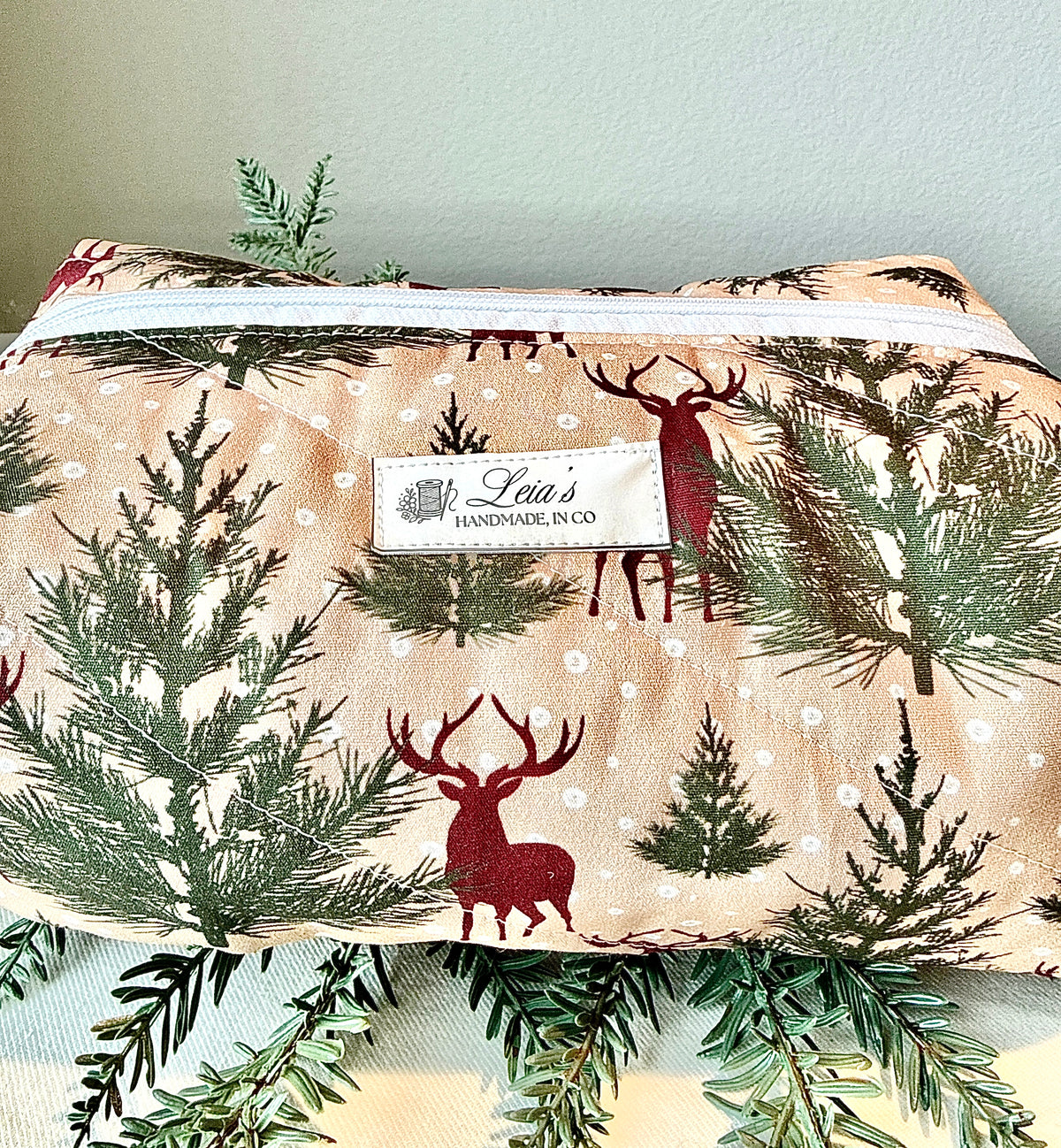 Close-up of the Winter Stag bag’s burgundy deer and evergreen print, white zipper, quilting, and Leia’s label.⠋
