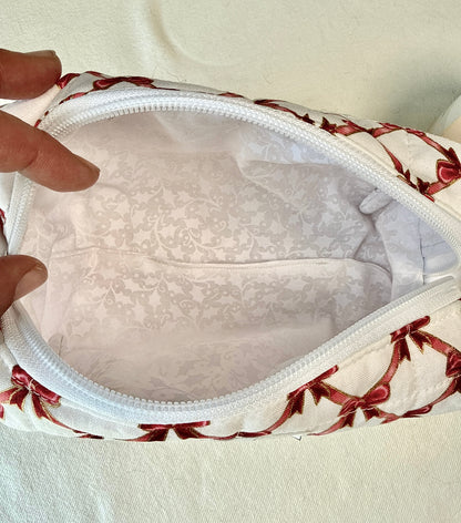 A close-up of a zippered fabric pouch opened by a hand, showing a white interior with a subtle damask pattern and a white zipper. The exterior fabric has a red bow and vine motif on a white background, set against a light fabric backdrop.