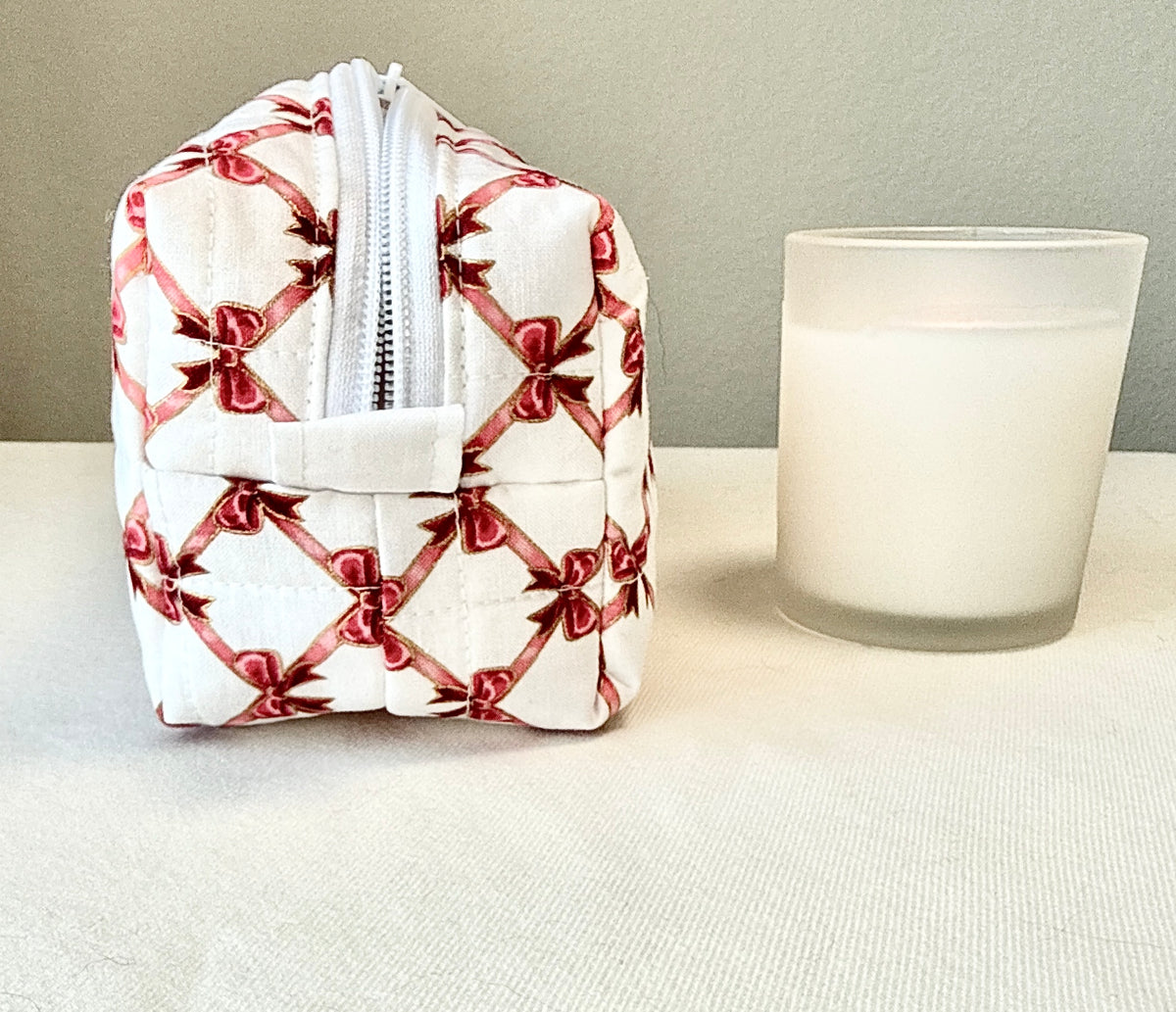 A small quilted white zippered pouch with a red bow and ribbon pattern, shown next to a frosted glass candle on a light fabric surface against a neutral wall.