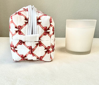A small quilted white zippered pouch with a red bow and ribbon pattern, shown next to a frosted glass candle on a light fabric surface against a neutral wall.